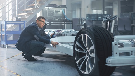 Engineer with Glasses Works on a Tablet Computer Next to an Electric Car Chassis Prototype with Wheels, Batteries and Engine in a High Tech Development Laboratory.